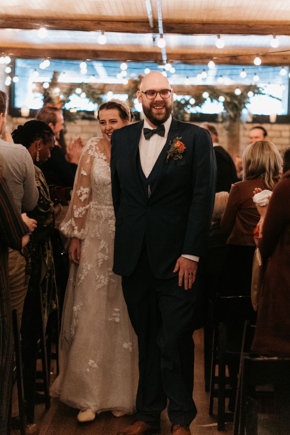 Laughing groom and smiling bride exit wedding ceremony through aisle of cheering guests in rustic venue.