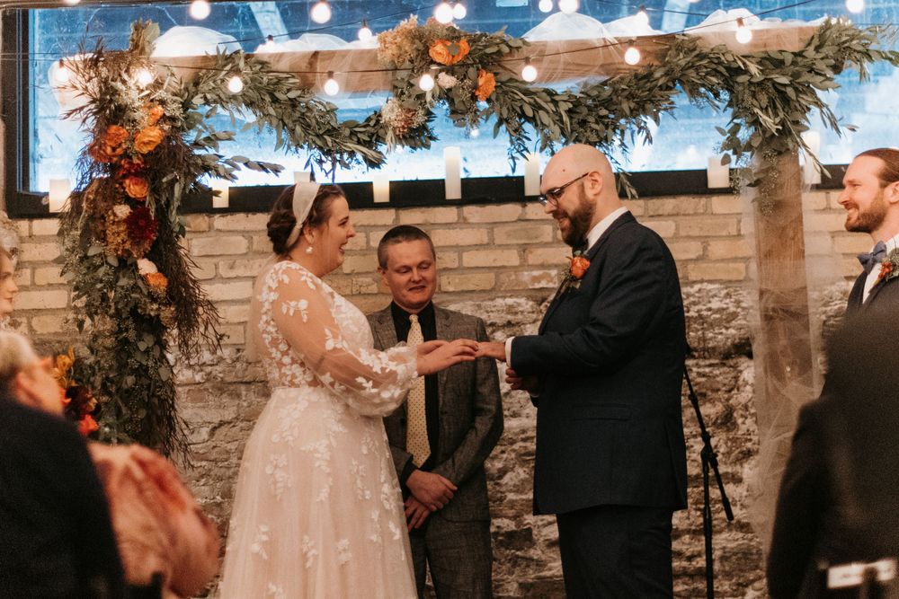 Bride places wedding ring on groom's finger during indoor ceremony with autumn floral arch and string lights.