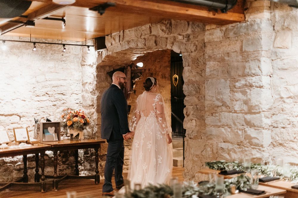 Newlywed couple walks hand in hand toward stone archway exit in a rustic wedding venue.