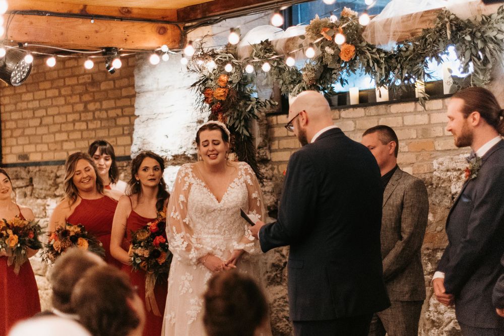 Bride smiles as groom holds her hands during emotional indoor wedding ceremony.