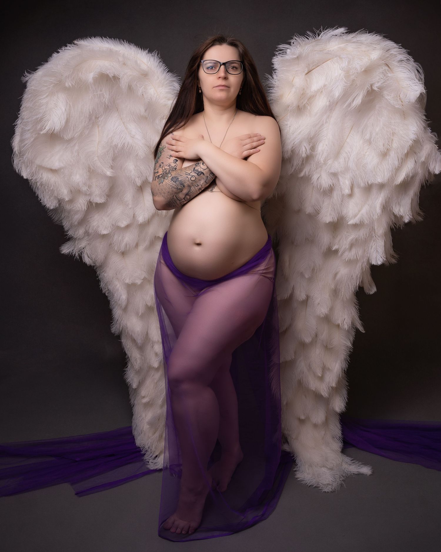Artistic maternity photo with large white angel wings and flowing purple fabric.