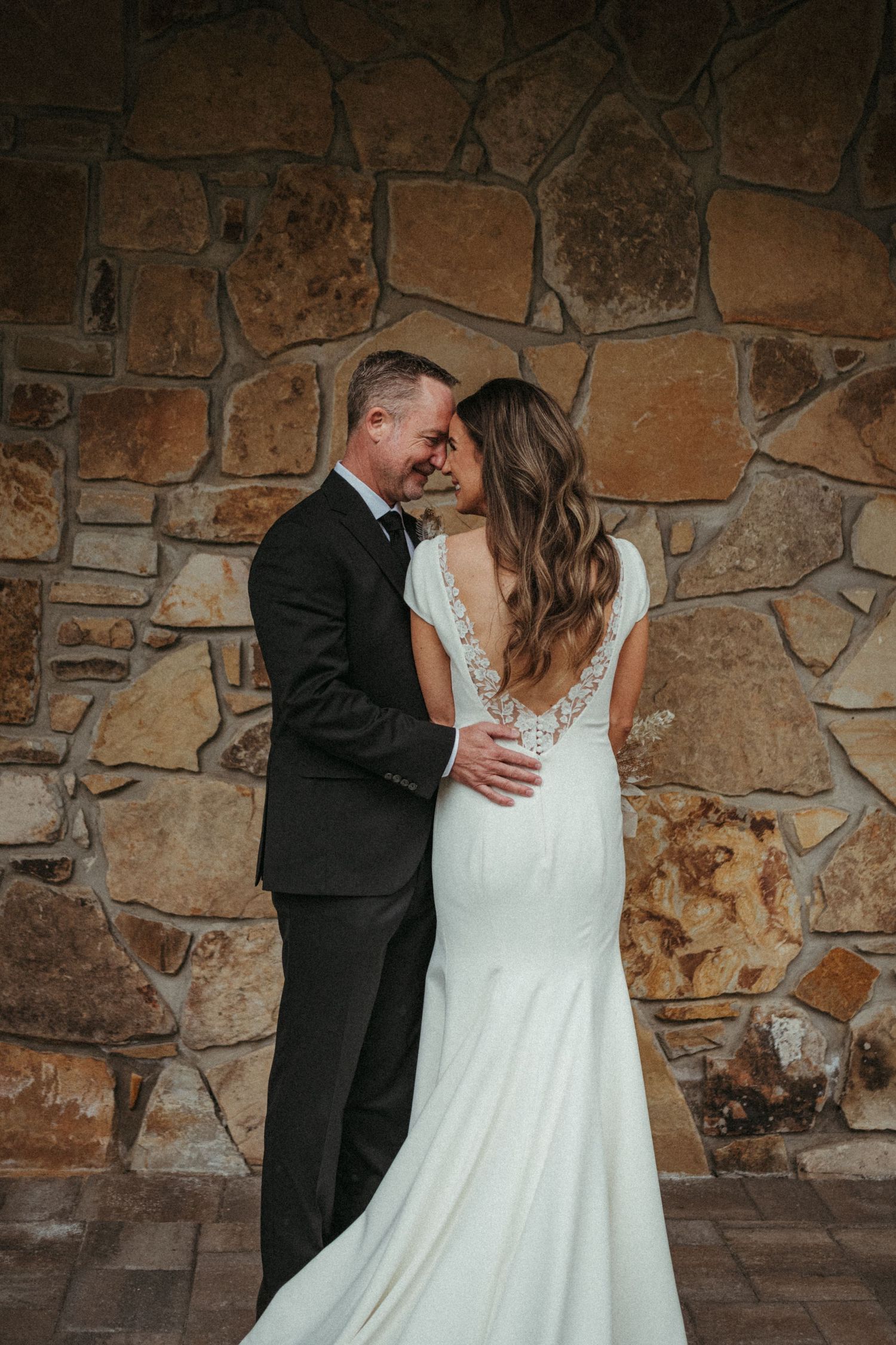 Intimate wedding couple poses against rustic stone wall backdrop.