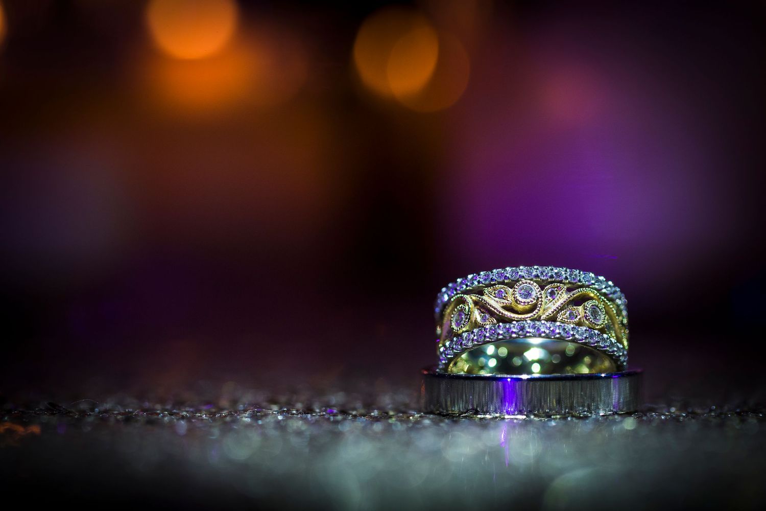 Ornate diamond wedding ring with intricate details photographed on dark surface with bokeh lighting effects.
