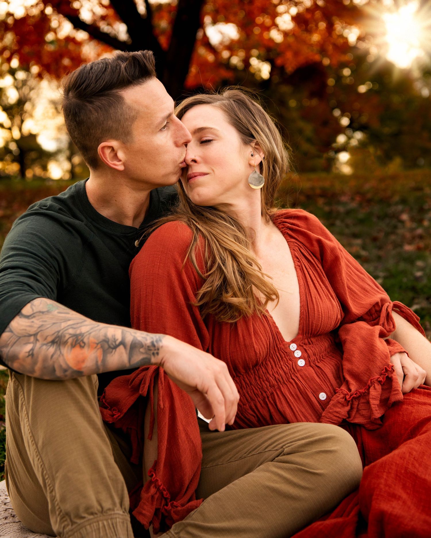 A tattooed man kisses a woman in a red dress as they sit together beneath autumn foliage.