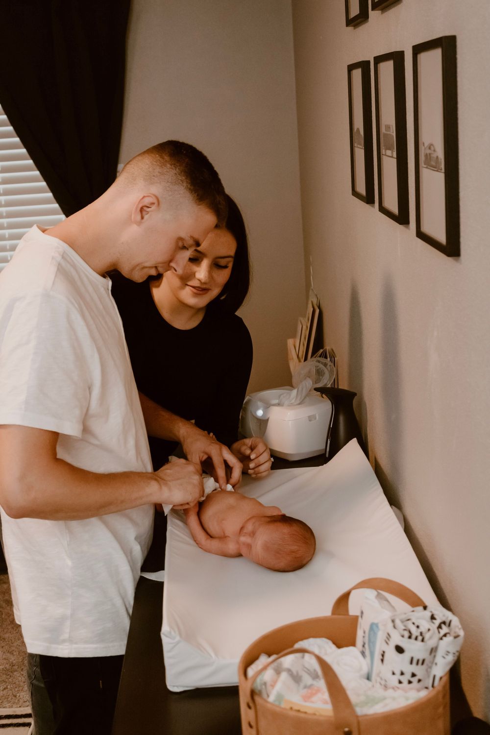 Parents caring for infant on white changing table with nursery supplies nearby.