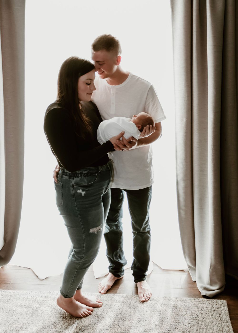 Couple stands by window curtains holding their newborn baby in a heartwarming family moment.