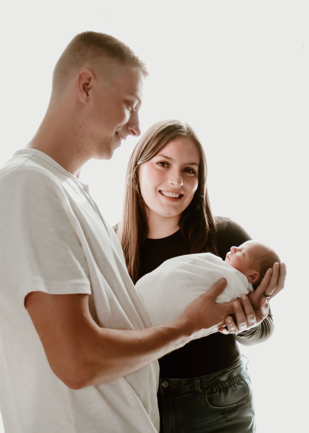 Parents share a tender moment while holding their newborn baby wrapped in white.