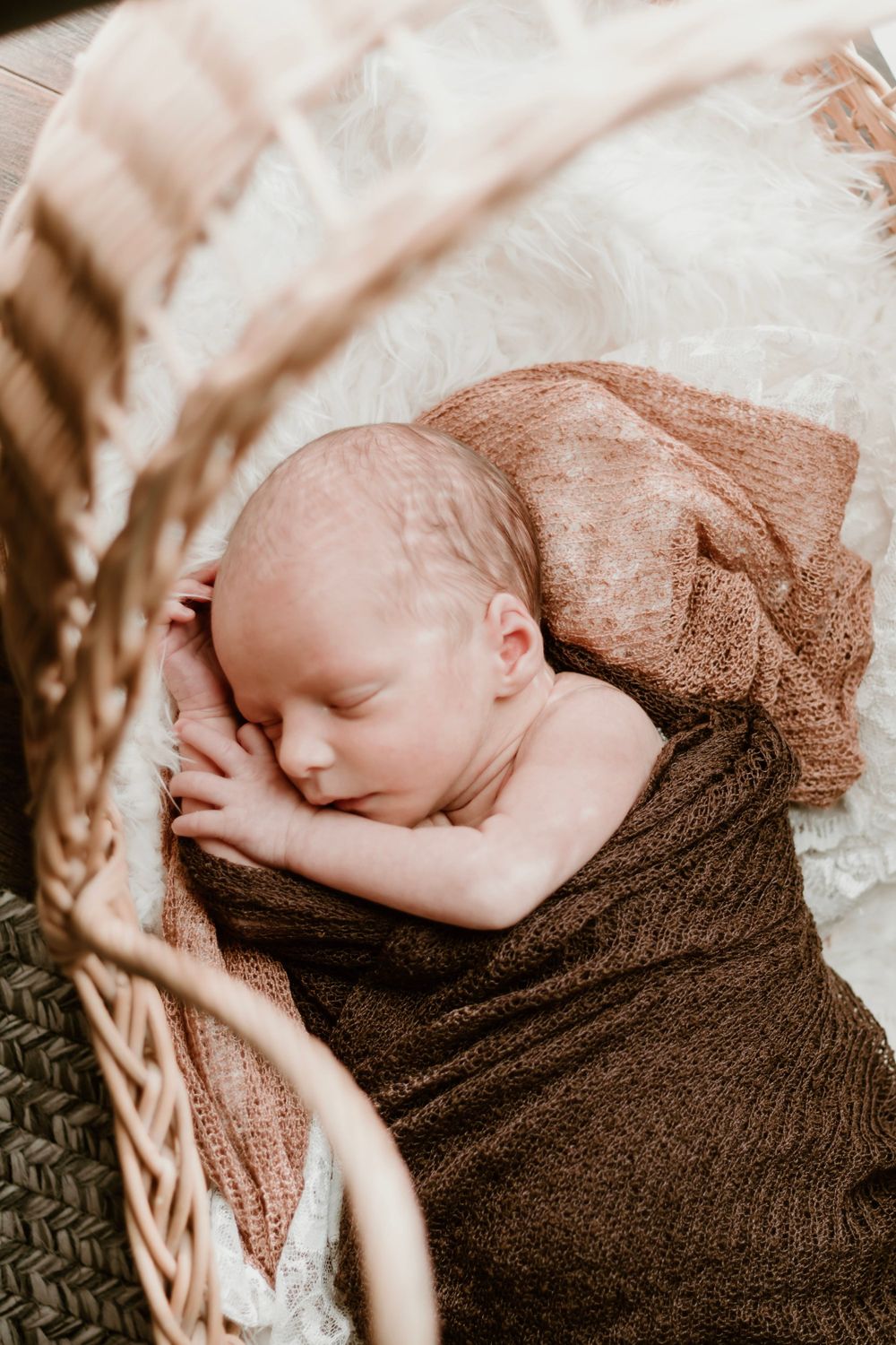 Sleeping newborn baby wrapped in brown knit blanket resting in wicker basket.