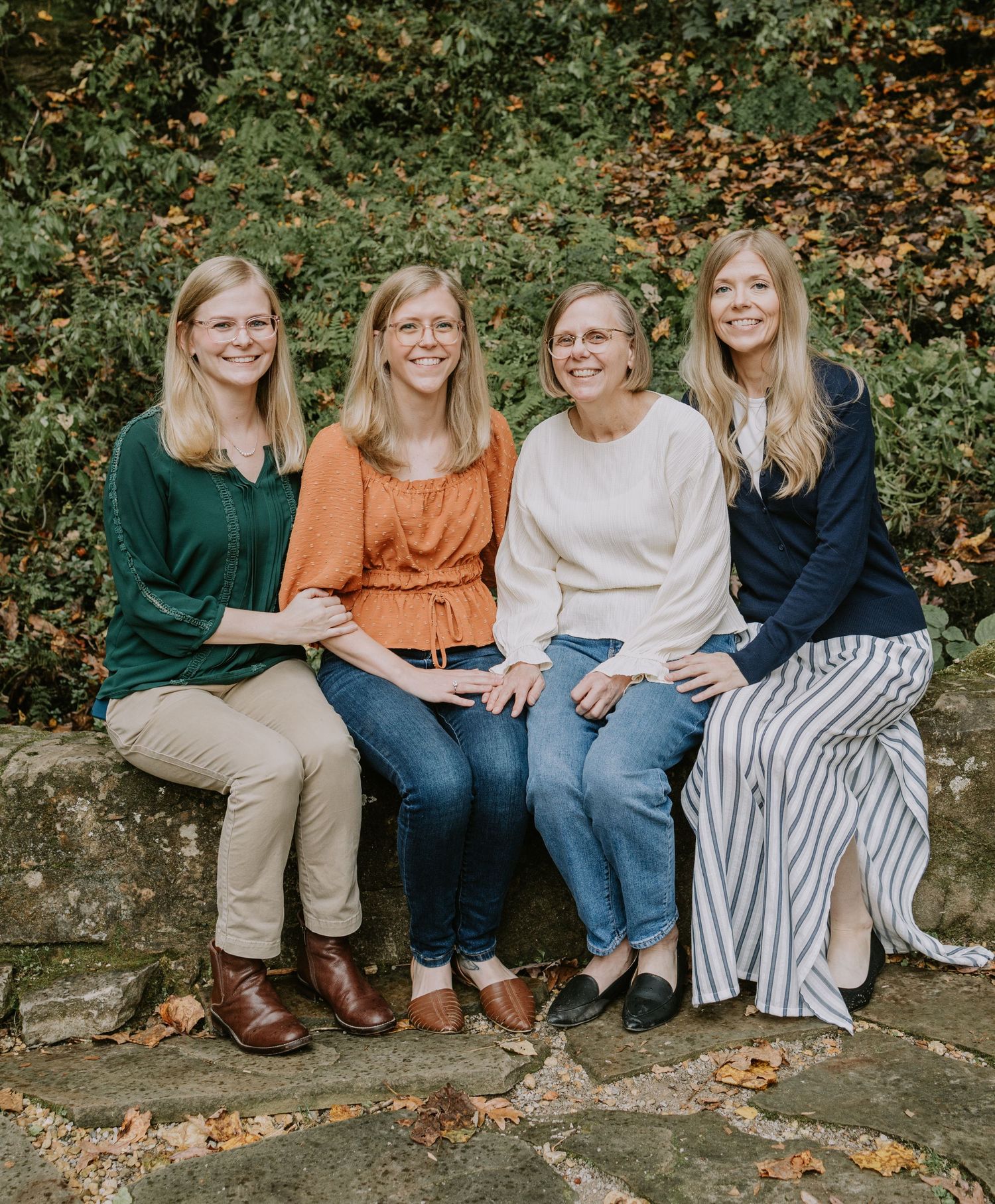 Four women in coordinating sweaters sit together on a stone wall during fall.