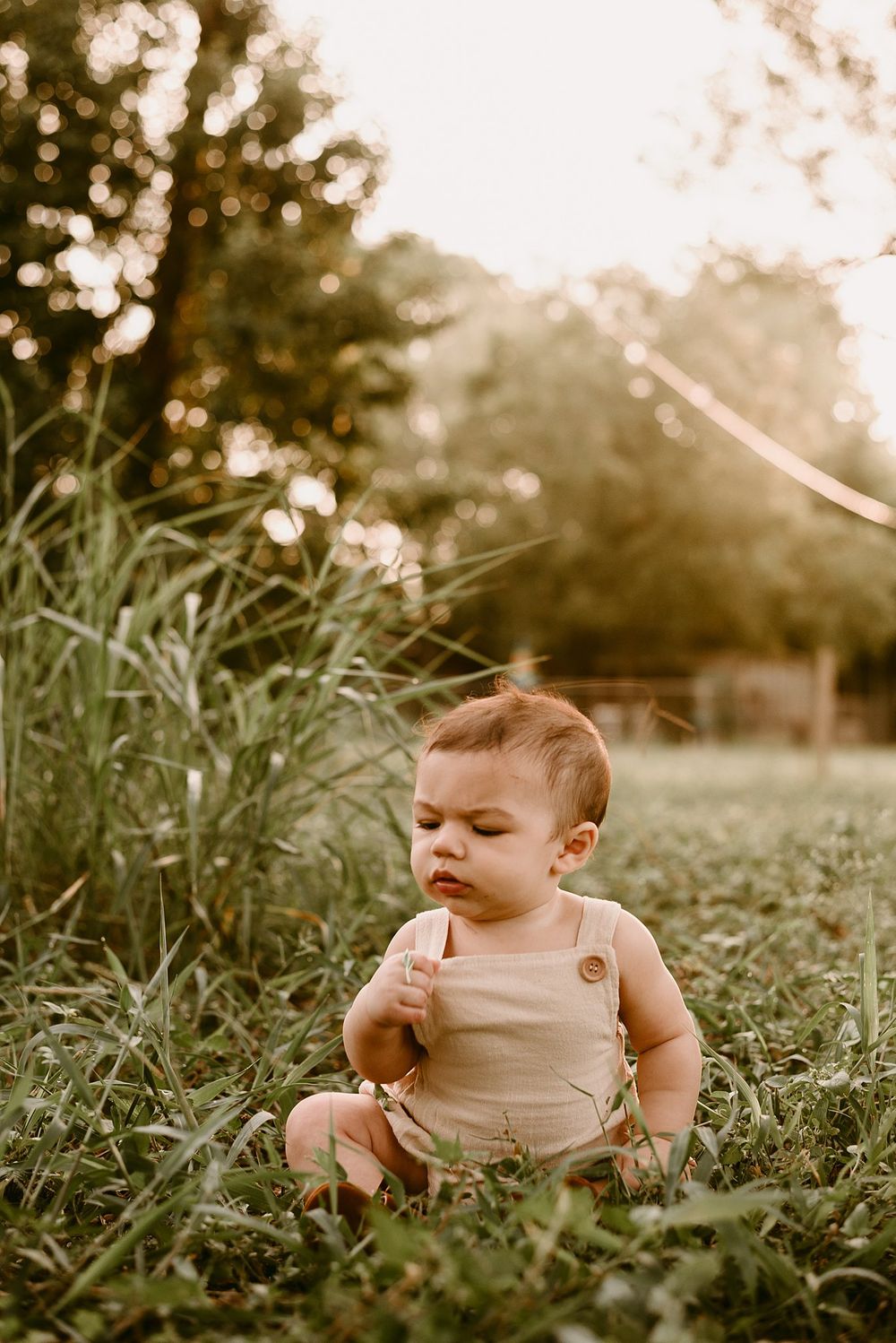 Baby in beige romper sits thoughtfully in green grass during golden hour sunset.