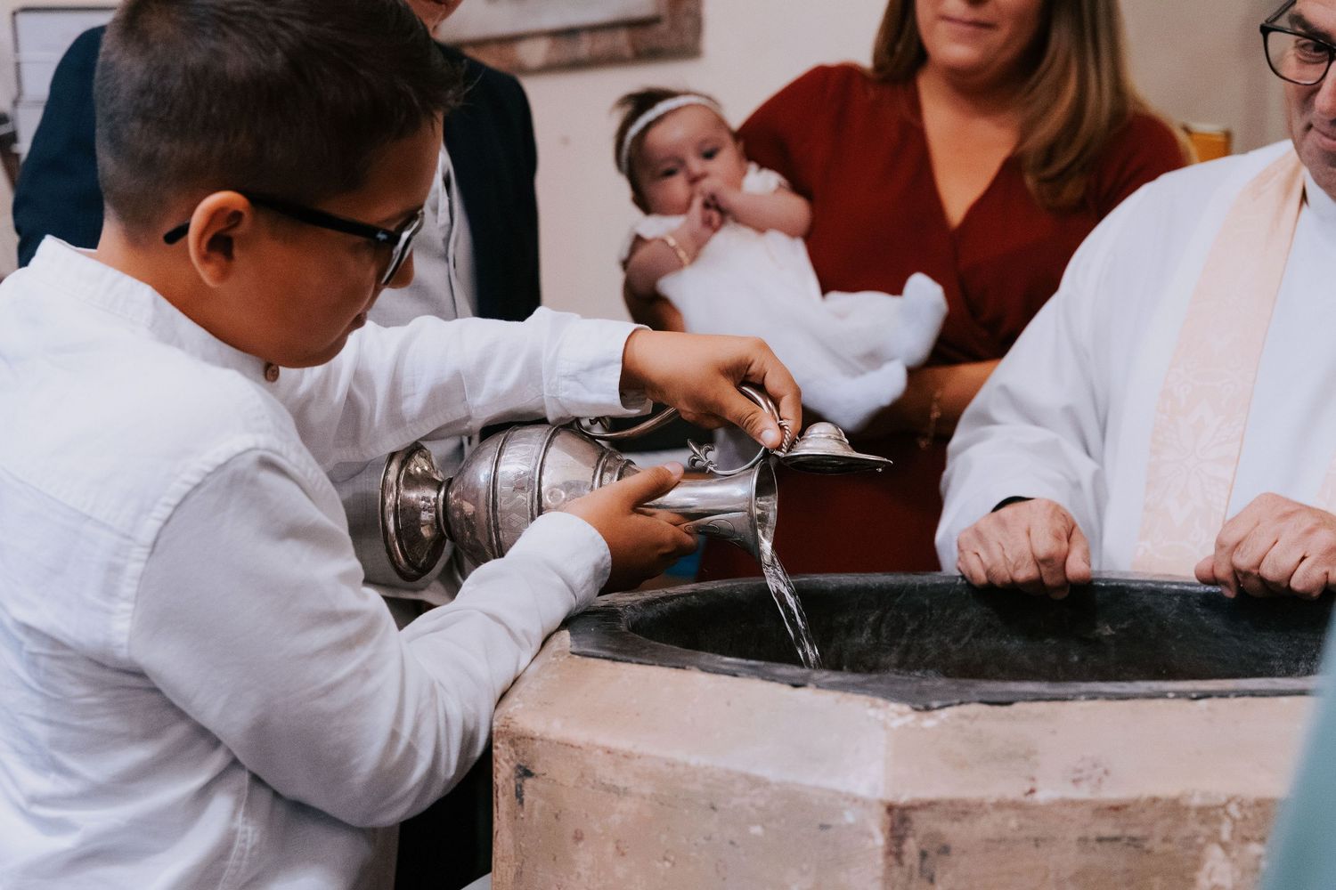 A child in white clothing leans over a baptismal font during a religious ceremony.