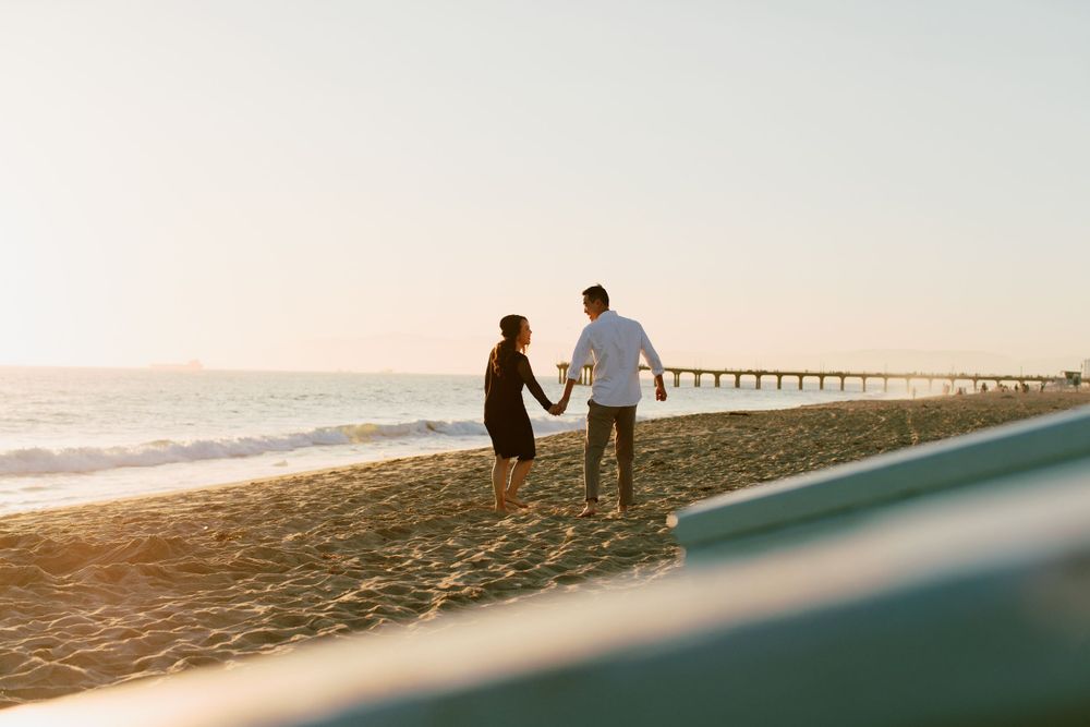 Two silhouettes walk along the beach at sunset with a pier in the distance.
