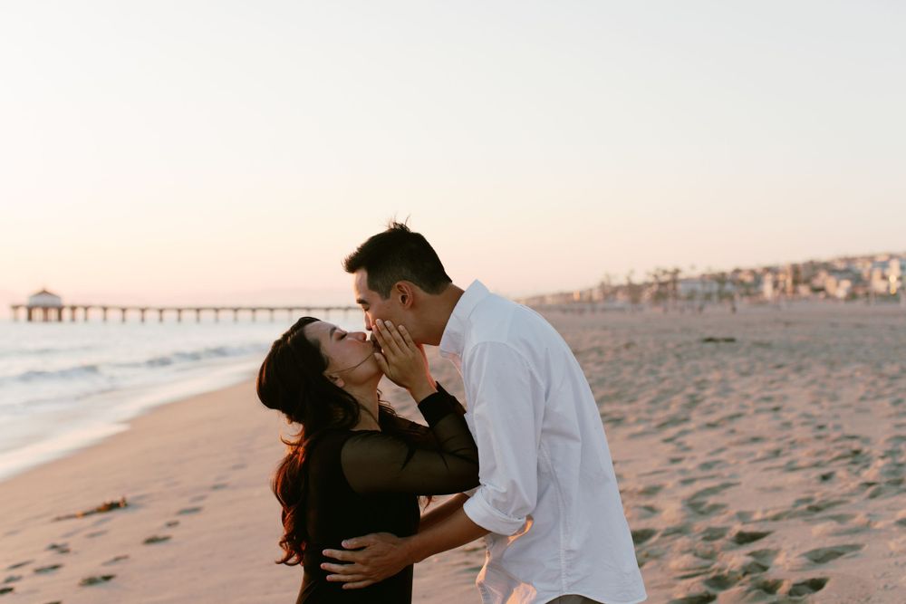 A romantic couple shares an intimate moment on a beach at sunset with a pier in the background.
