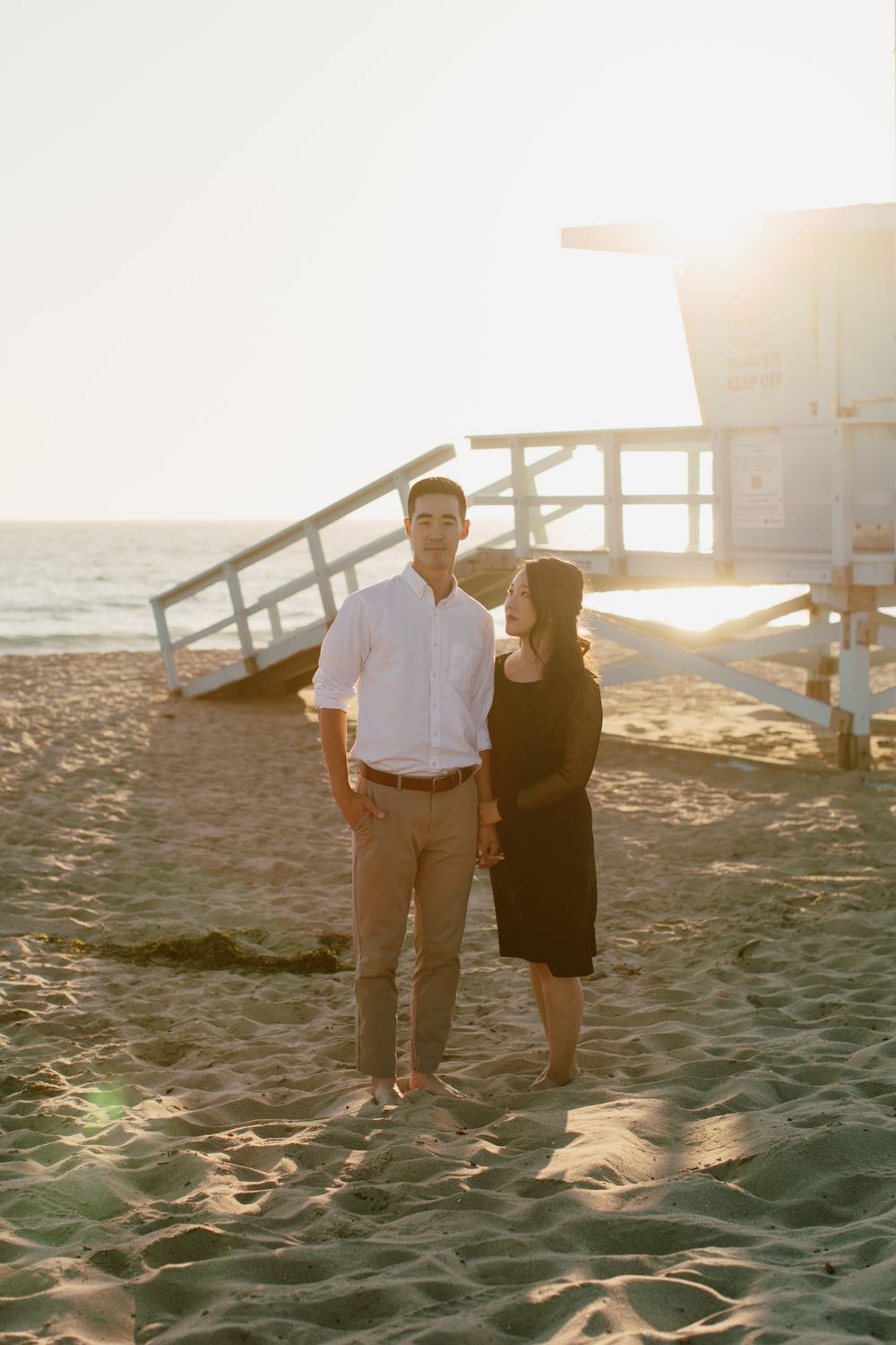 Couple standing together on beach with lifeguard tower during sunset.
