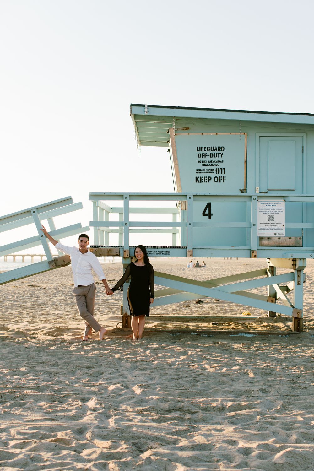 A couple stands next to a blue lifeguard tower numbered 4 on a sandy beach at sunset.