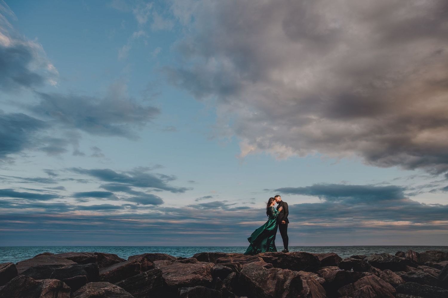 Dramatic coastal engagement photo with couple standing on rocky shoreline under cloudy skies.