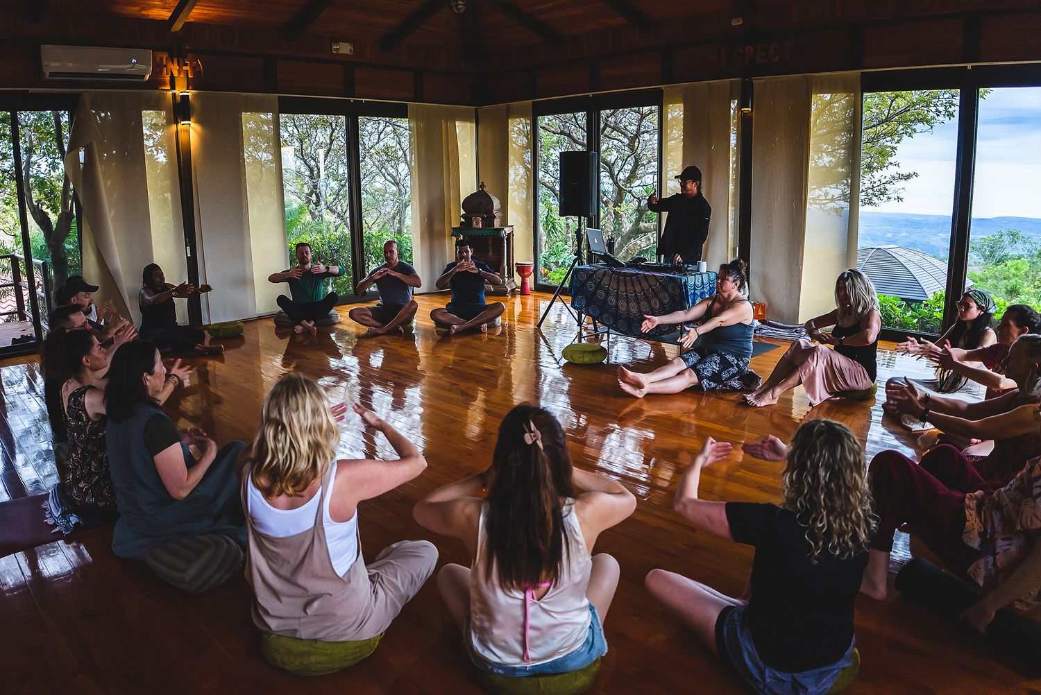 Large group participating in yoga or meditation class in sunlit room with panoramic windows.