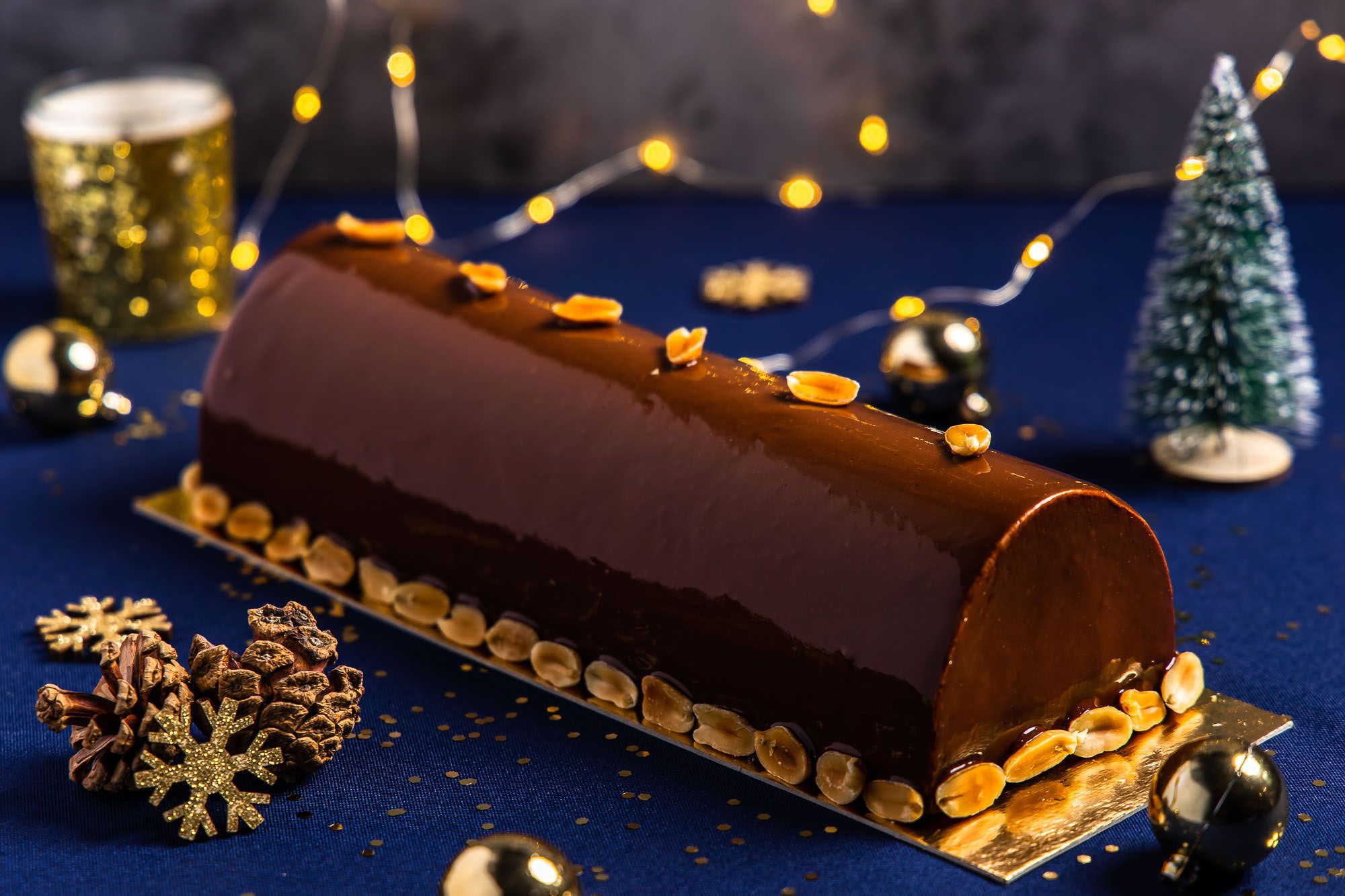 Elegant chocolate yule log cake decorated with gold accents and holiday lights on dark blue background.