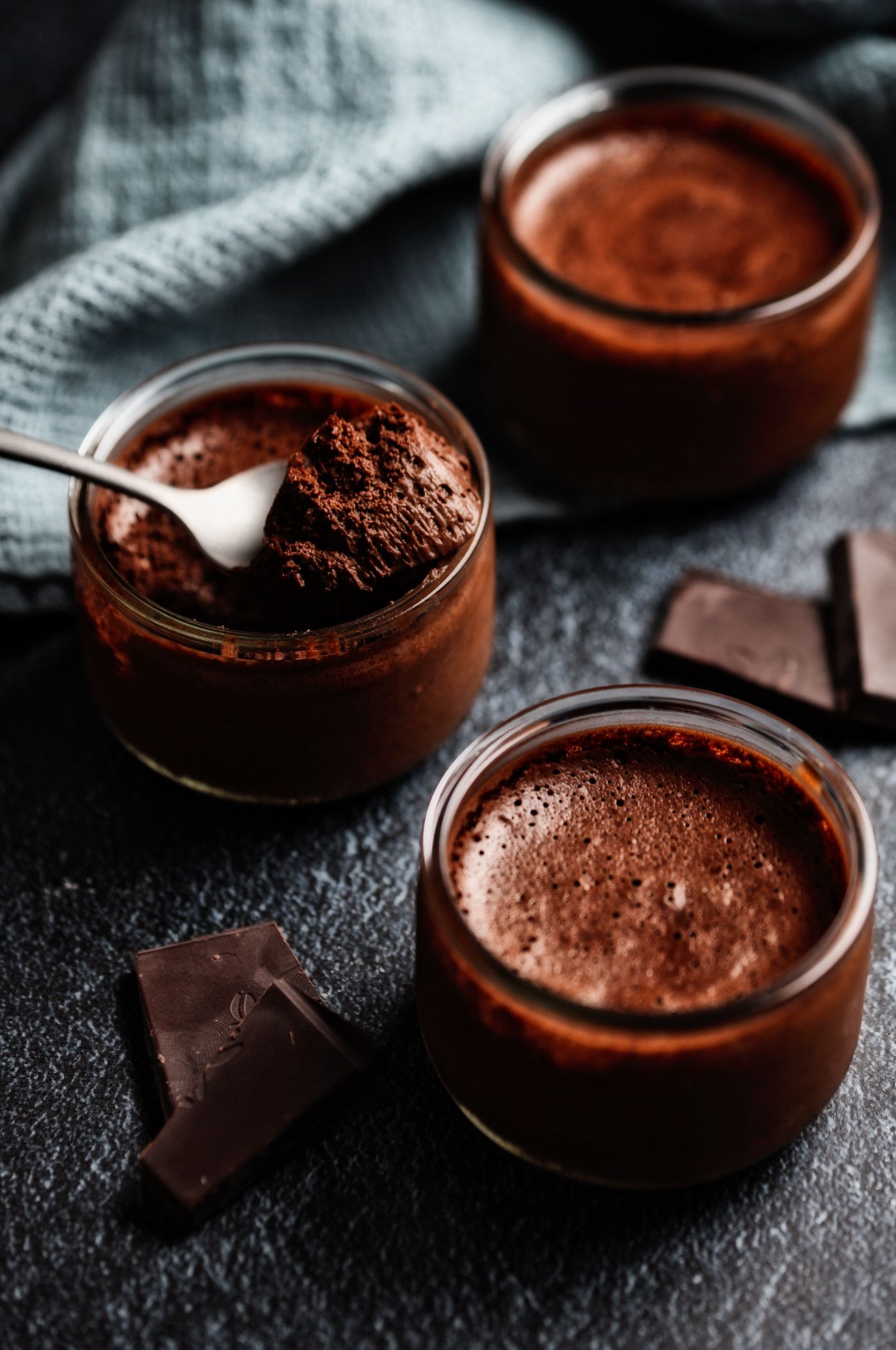 Rich chocolate mousse desserts served in glass bowls with dark chocolate pieces on a black surface.