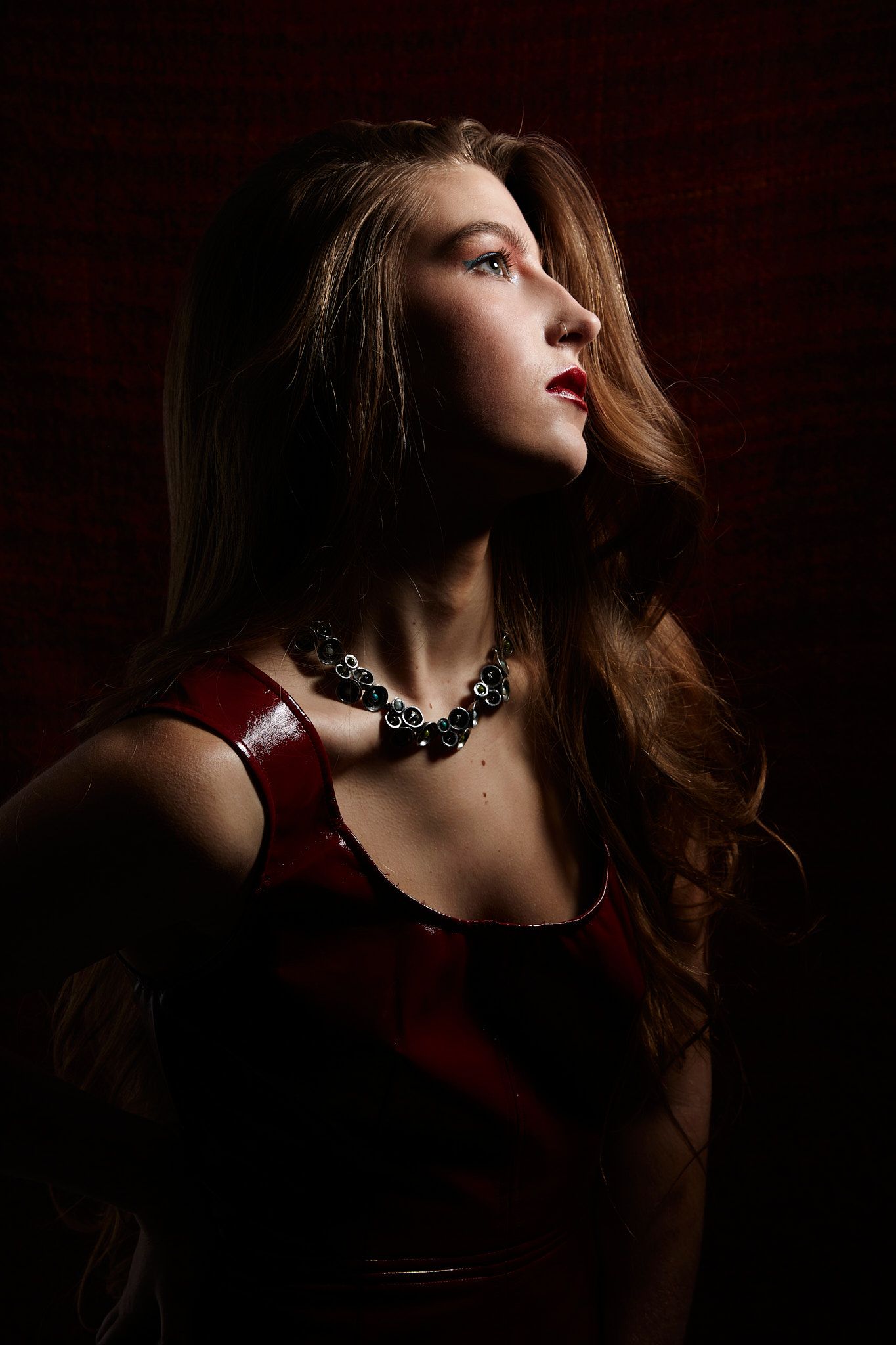 Dramatic portrait of someone in red dress with statement necklace against dark background, looking to side in chiaroscuro lighting.