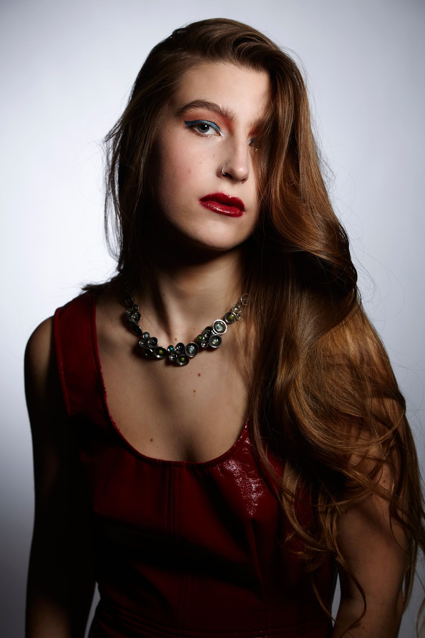 A dramatic fashion portrait featuring long brown hair, red lipstick, a burgundy top and an ornate statement necklace.