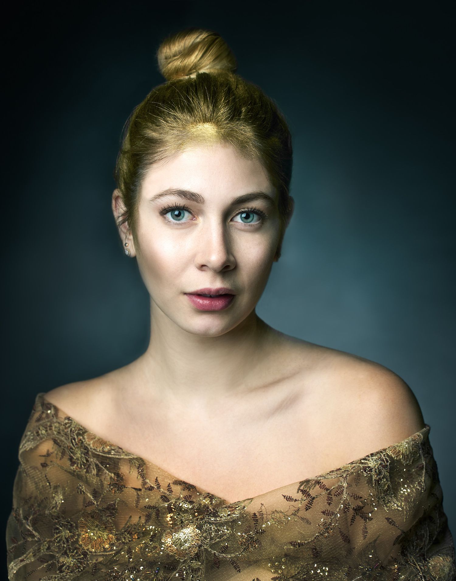 Portrait against dark background showing subject with blonde hair, bare shoulders, and wearing an ornate gold embroidered dress.