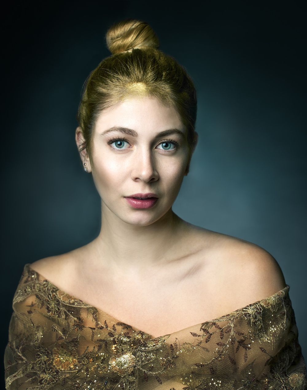 Portrait against dark background showing subject with blonde hair, bare shoulders, and wearing an ornate gold embroidered dress.