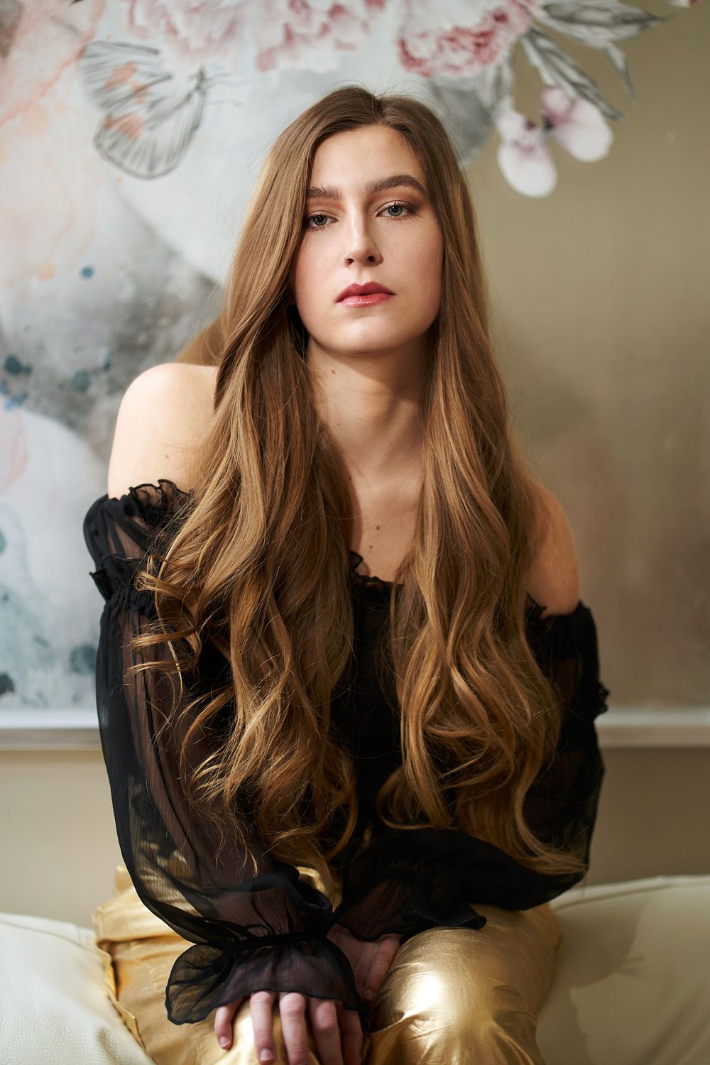 A portrait featuring long wavy brown hair, wearing a sheer black top against a soft artistic background.