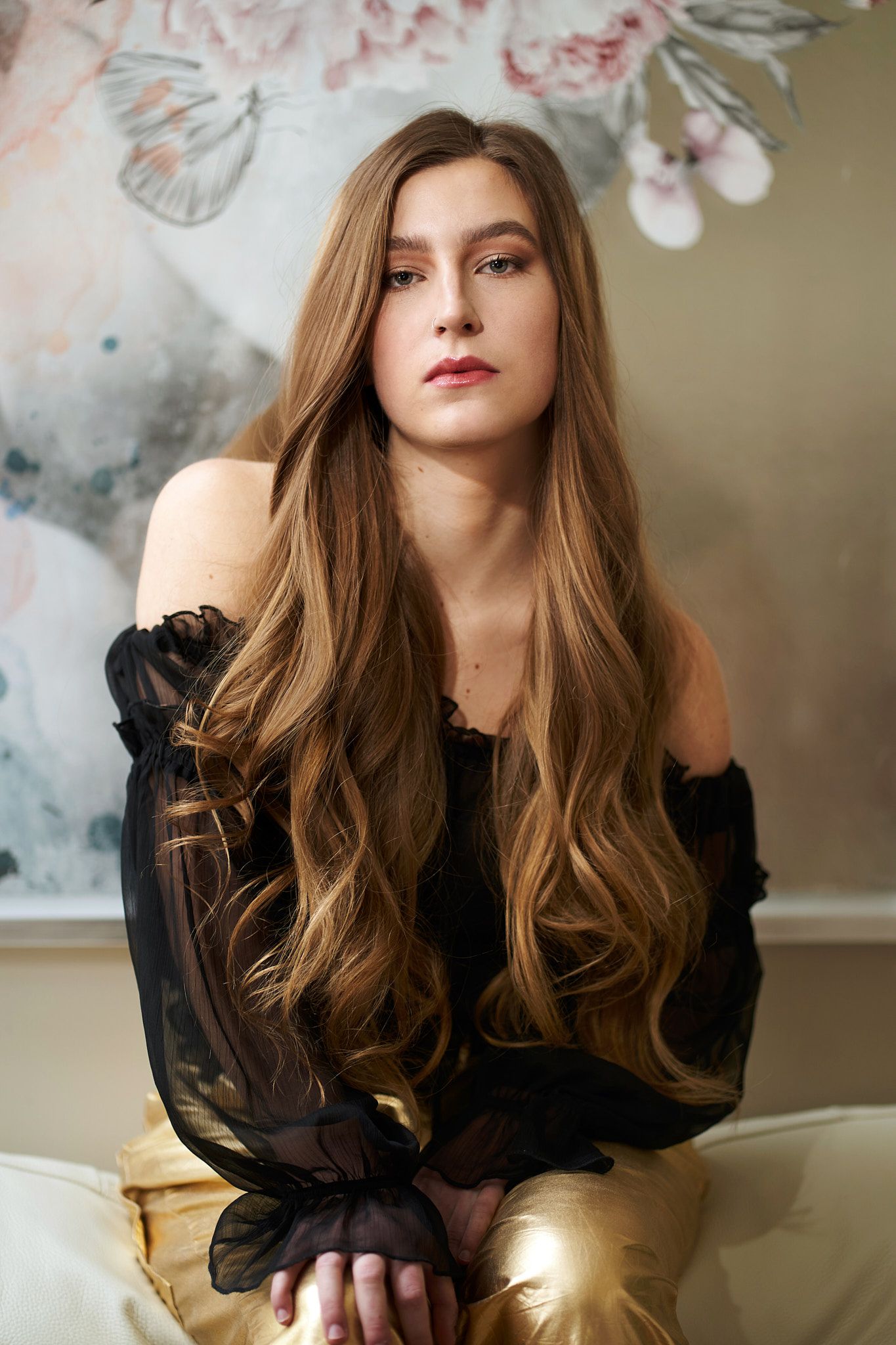 A portrait featuring long wavy brown hair, wearing a sheer black top against a soft artistic background.