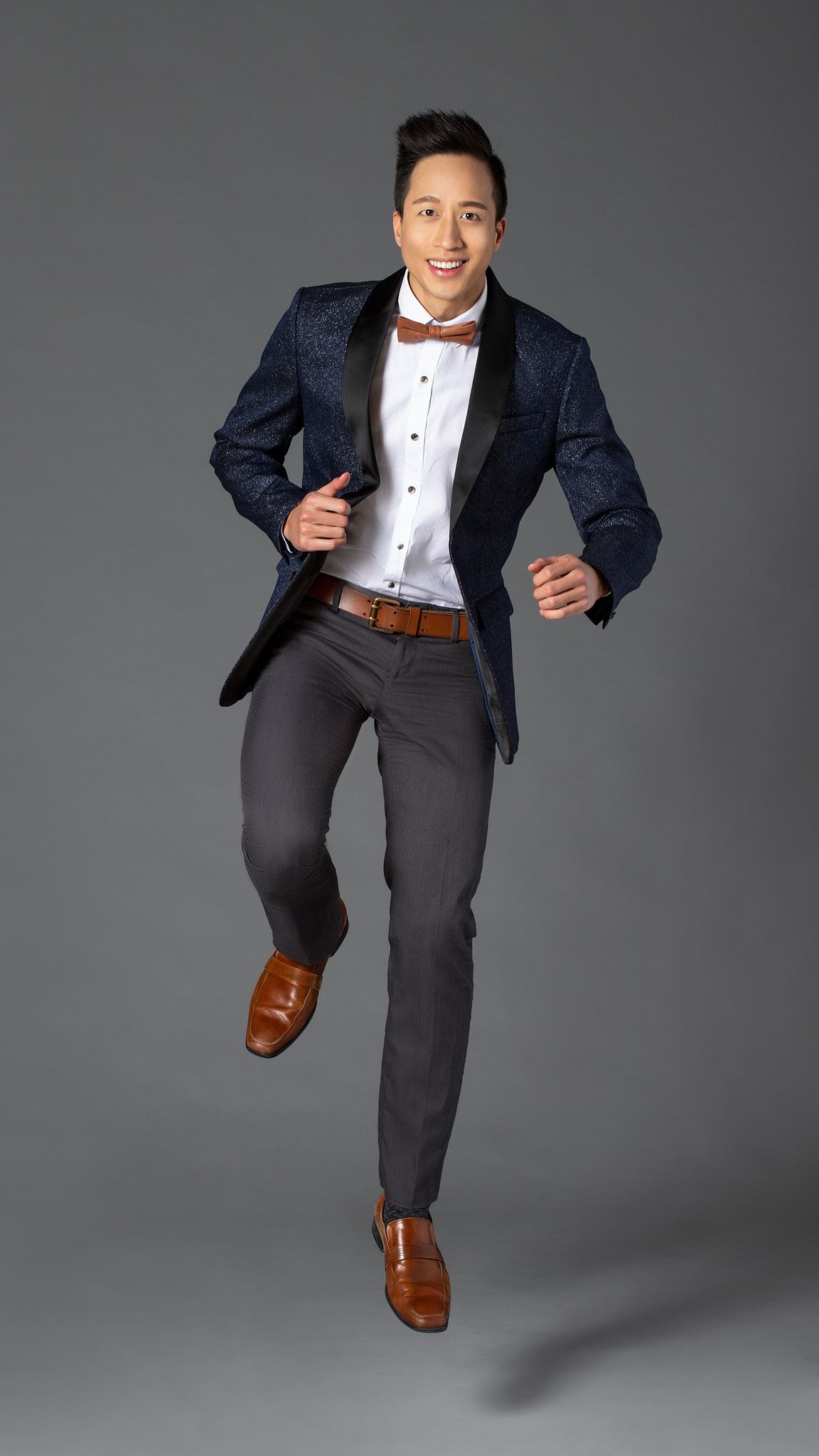 A person in a navy blazer and grey trousers jumps joyfully while wearing brown leather shoes against a grey backdrop for fashion portrait