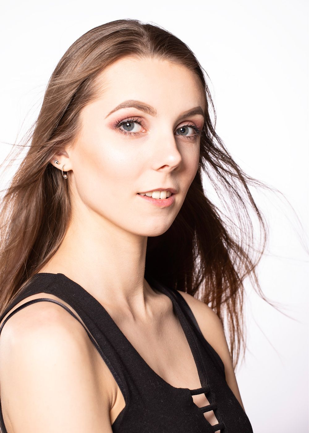 Studio portrait against white background showing long brown hair flowing in wind and wearing black sleeveless top.