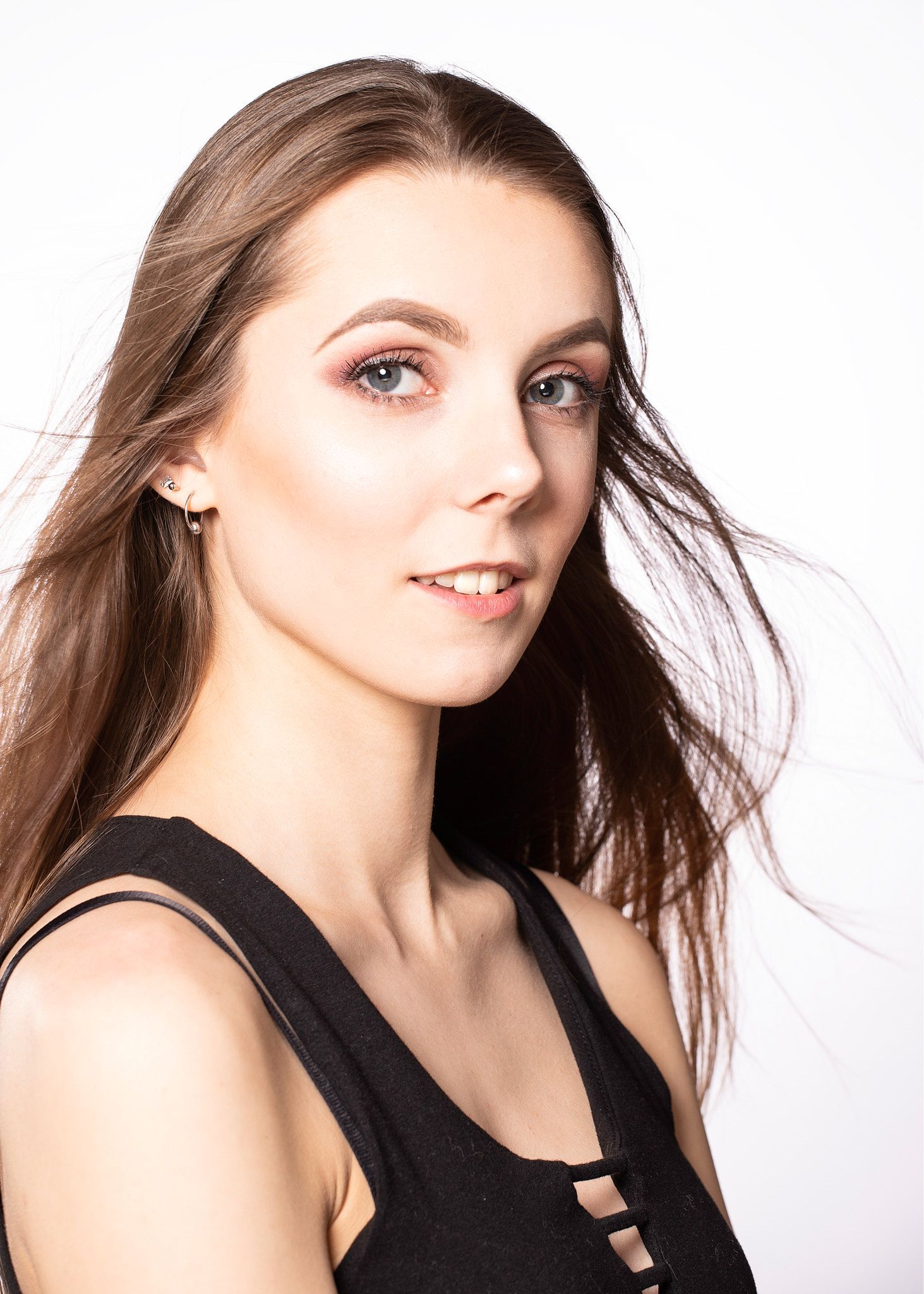 Studio portrait against white background showing long brown hair flowing in wind and wearing black sleeveless top.