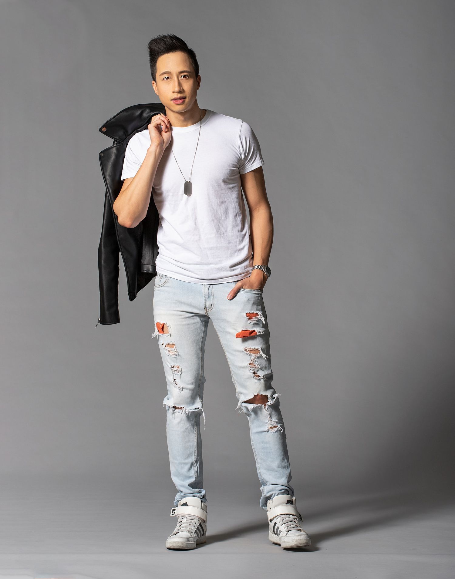 A person in distressed light jeans and white t-shirt poses casually with a black jacket over their shoulder for fashion shoot.