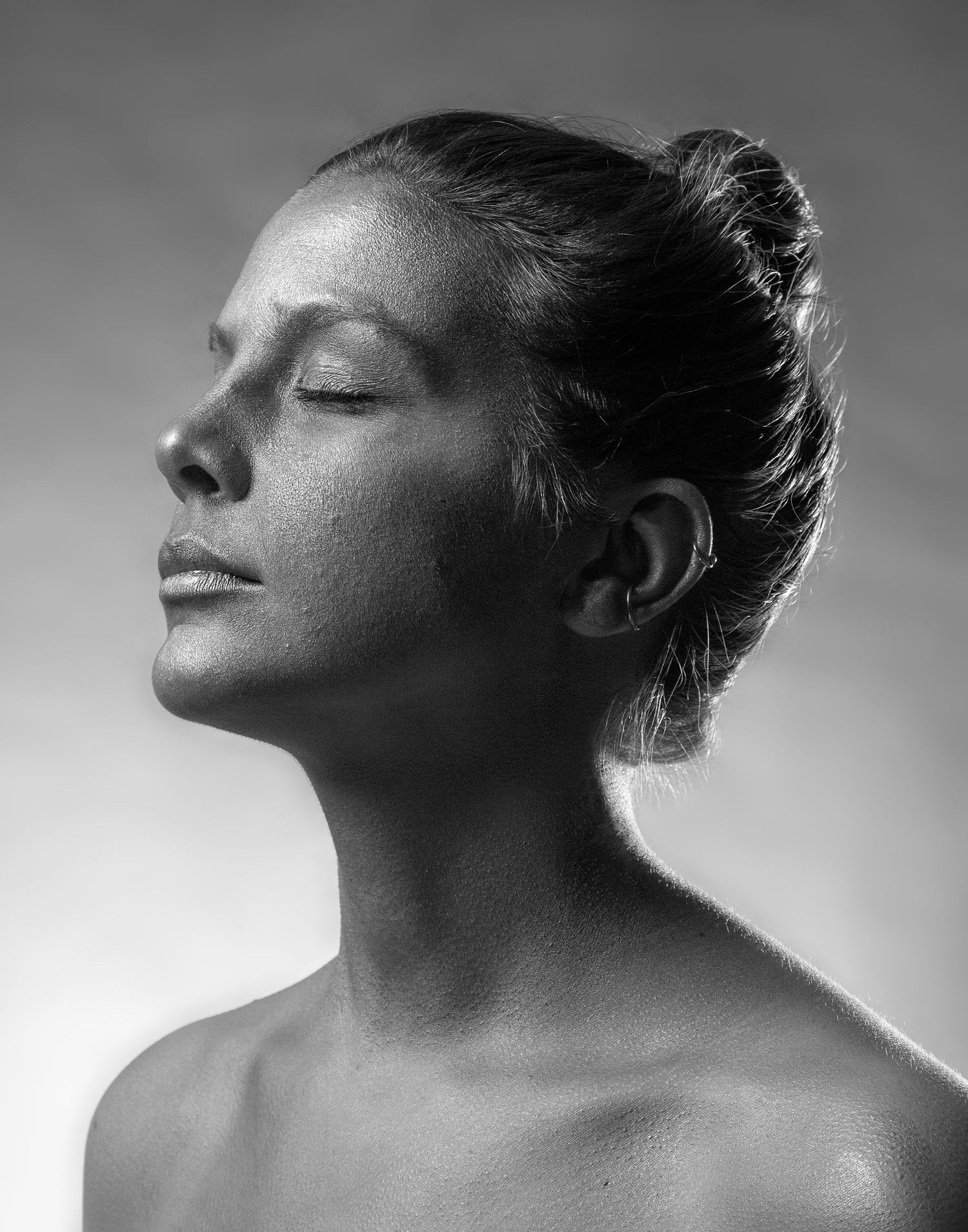 Artistic black and white profile portrait with eyes closed and hair in bun showing serene expression in soft lighting.