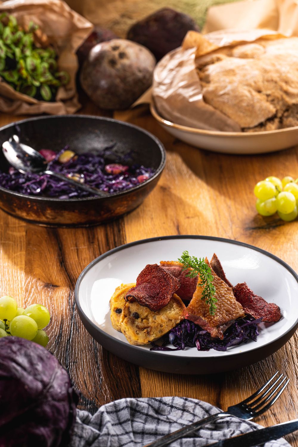 Elegant plated dish with red meat and purple cabbage served alongside fresh grapes and bread on rustic wooden table.