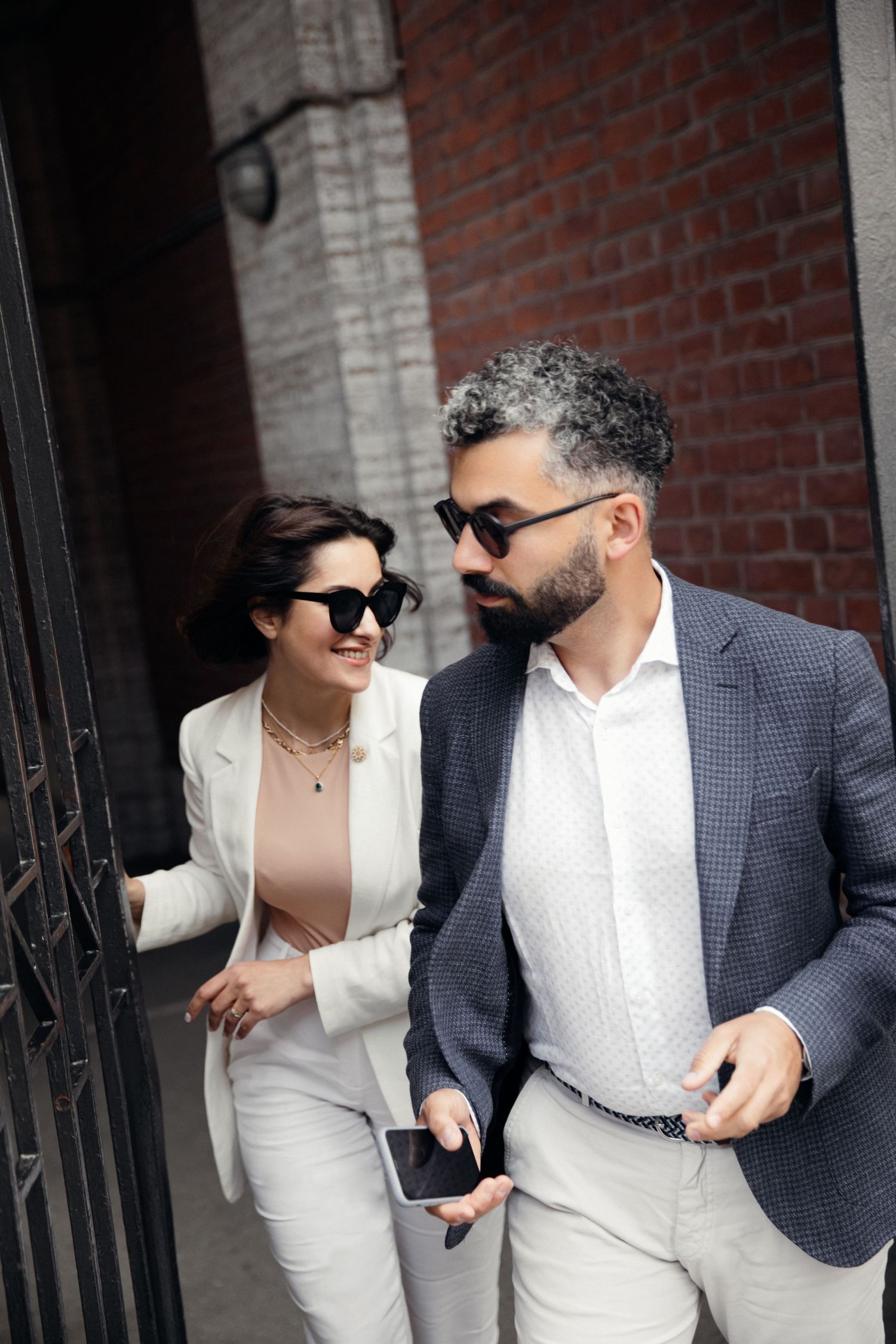 Stylish couple in formal attire walking through a doorway with brick wall background.