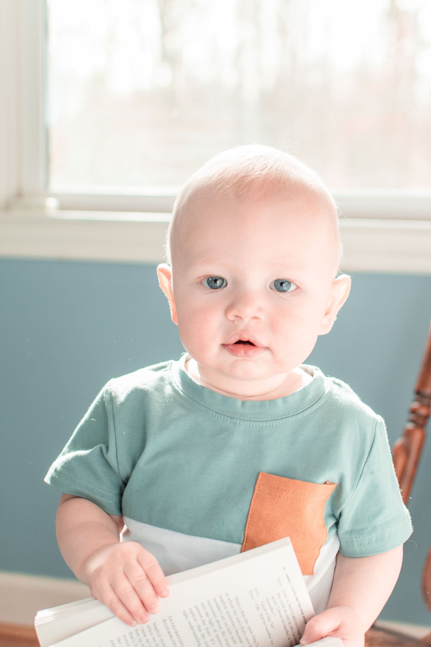 Infant in mint green t-shirt holding book near window in blue room.