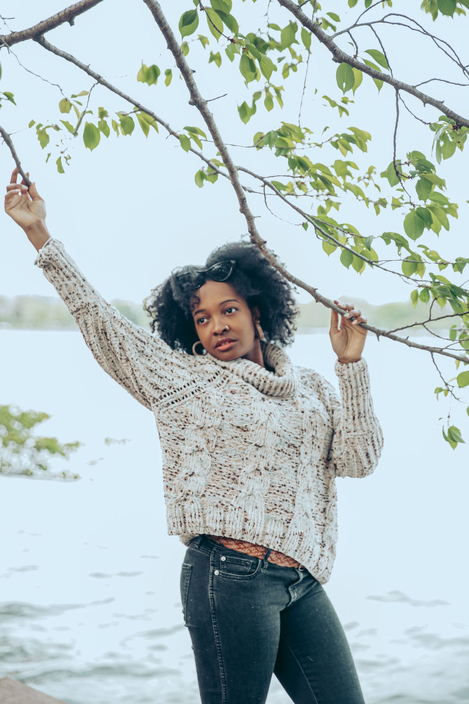 Young Black woman in chunky knit sweater and jeans posing under green tree branches near a lake.