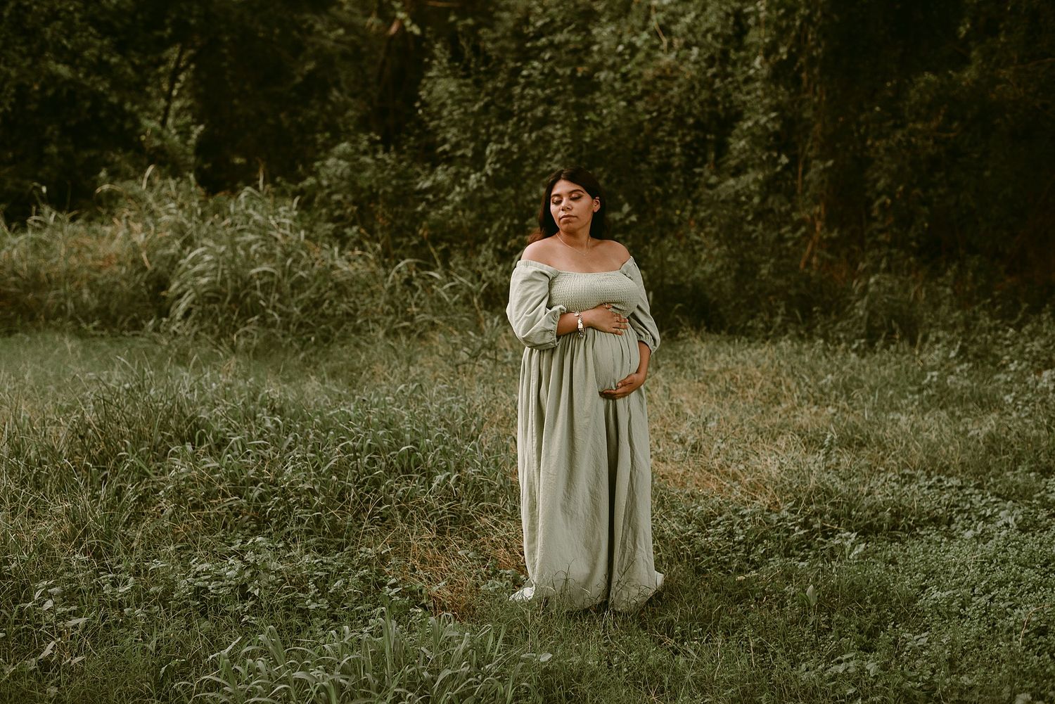 Figure in an ethereal white dress poses in a sunlit meadow surrounded by trees.