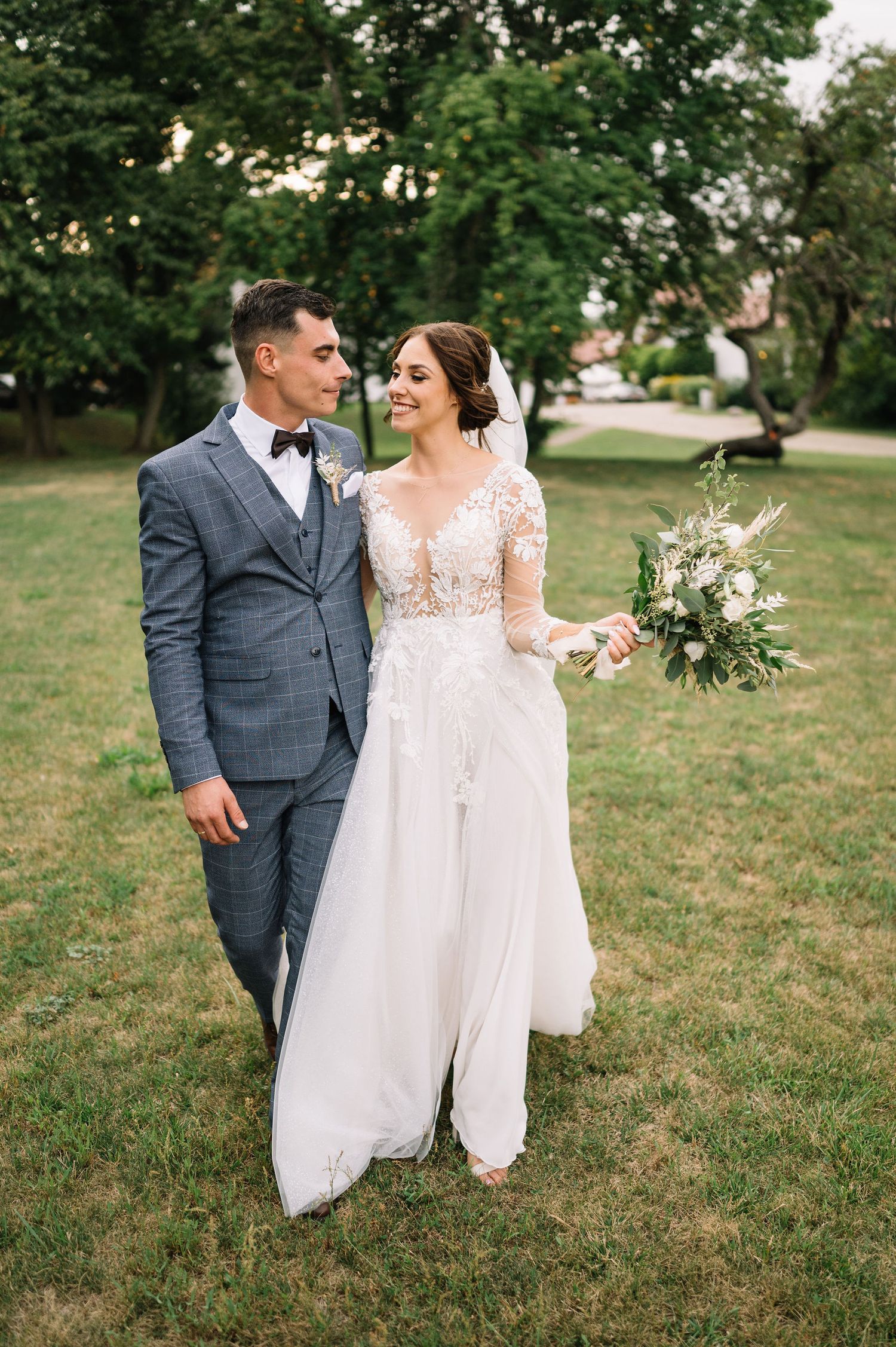 A wedding couple stands together on grass, bride in lace gown holding white bouquet.