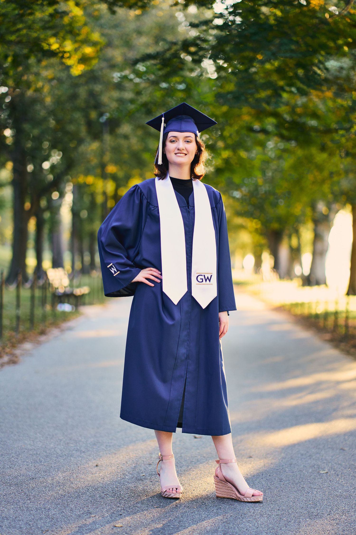 Graduate in navy blue cap and gown poses on tree-lined path in natural lighting.