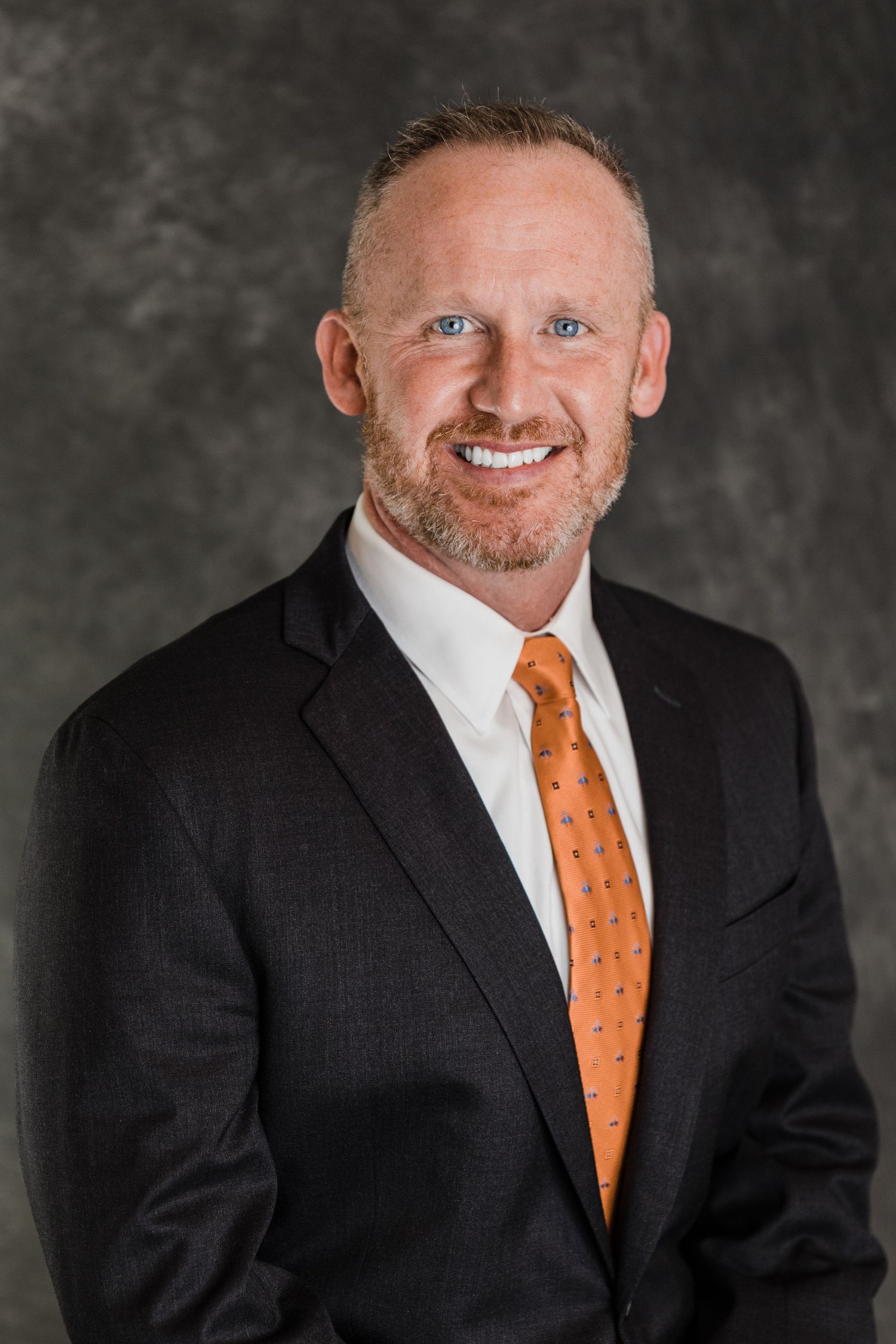 Professional headshot of businessman wearing dark suit with orange tie.