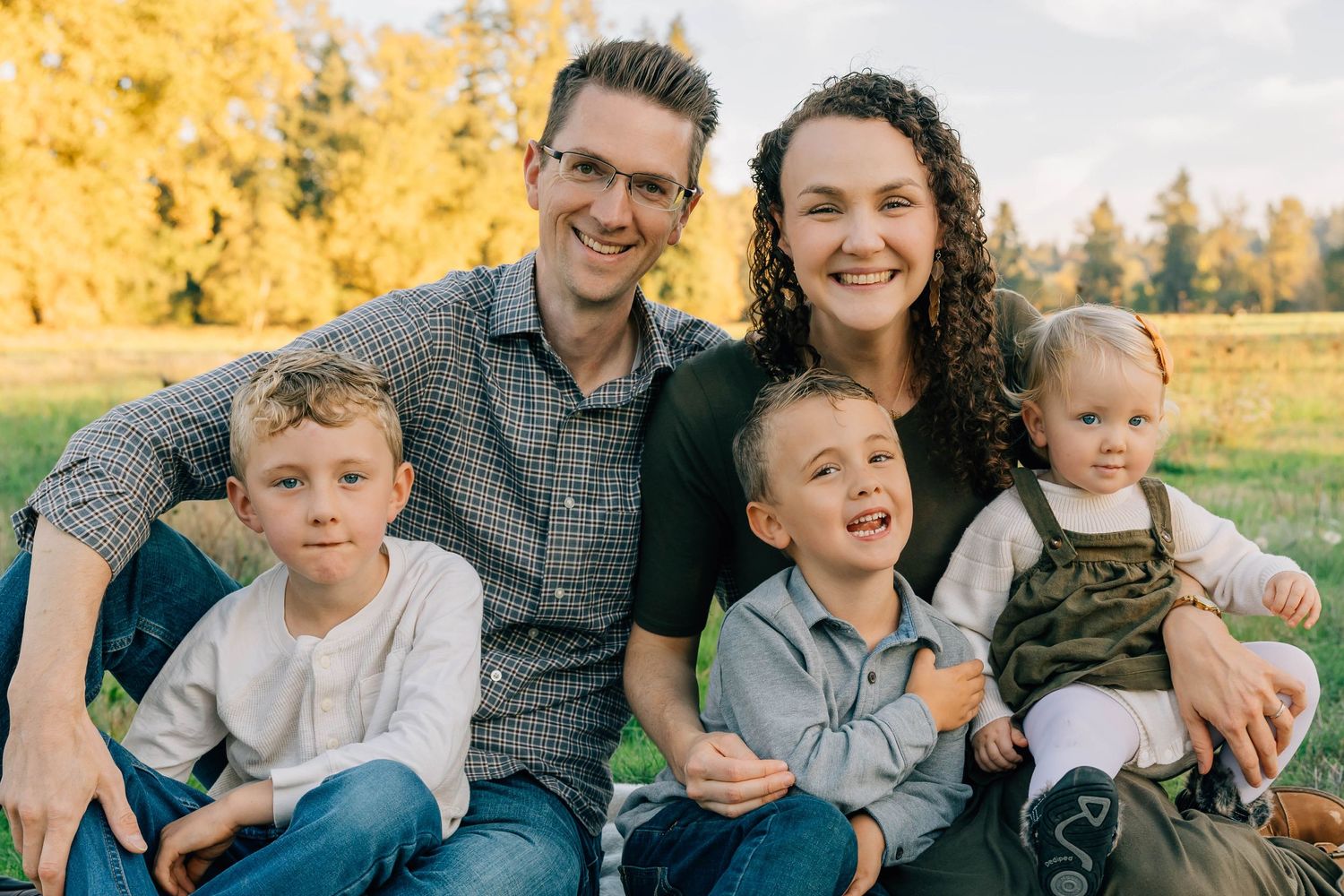 Happy family of five smiling outdoors in golden autumn light with green grass and trees.