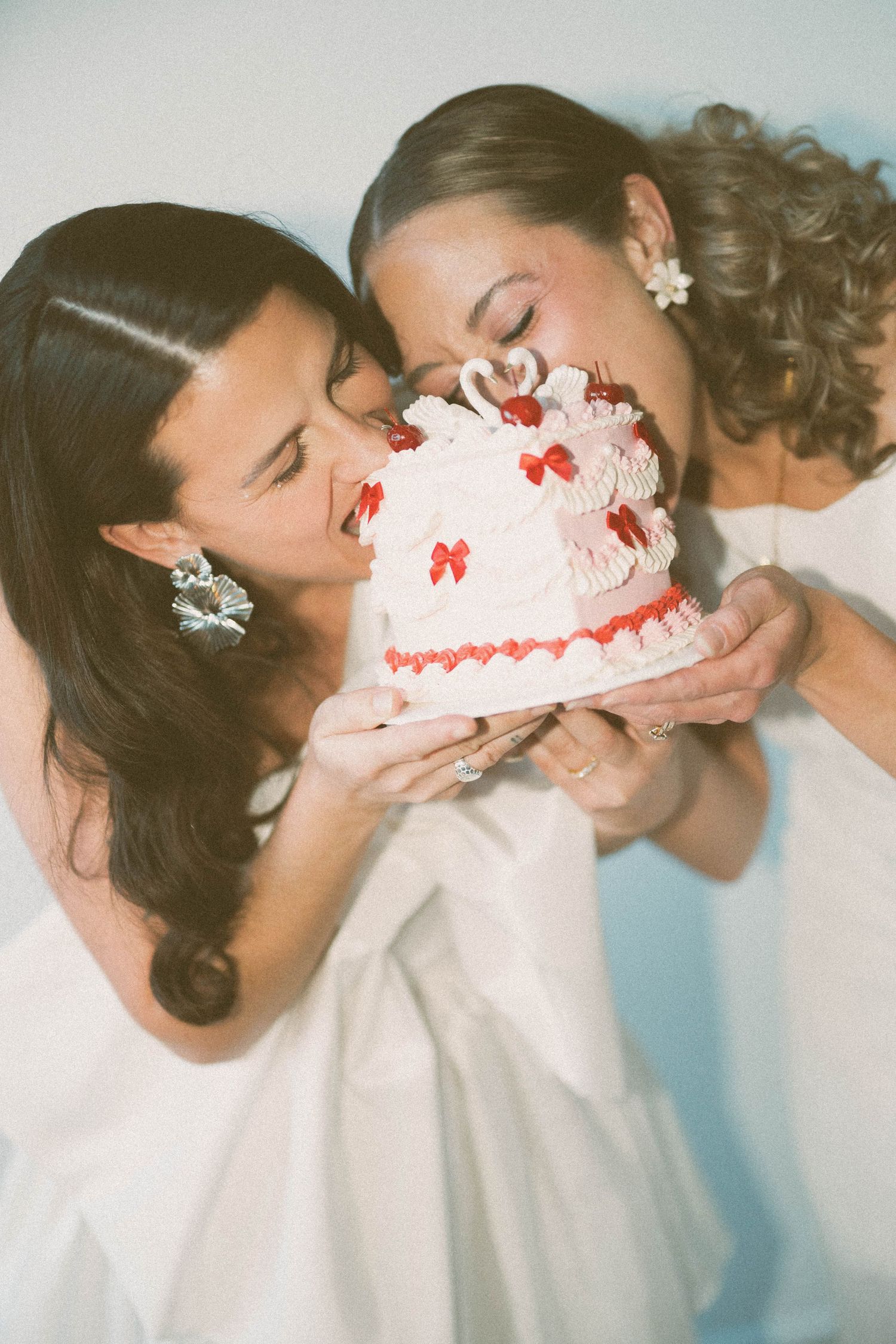 Two people playfully bite into a small white wedding cake with red decorations.