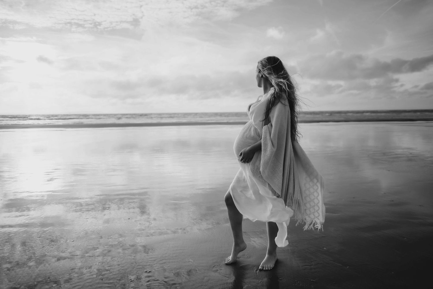 A flowing dress catches the wind as someone stands on a reflective beach at sunset in black and white.