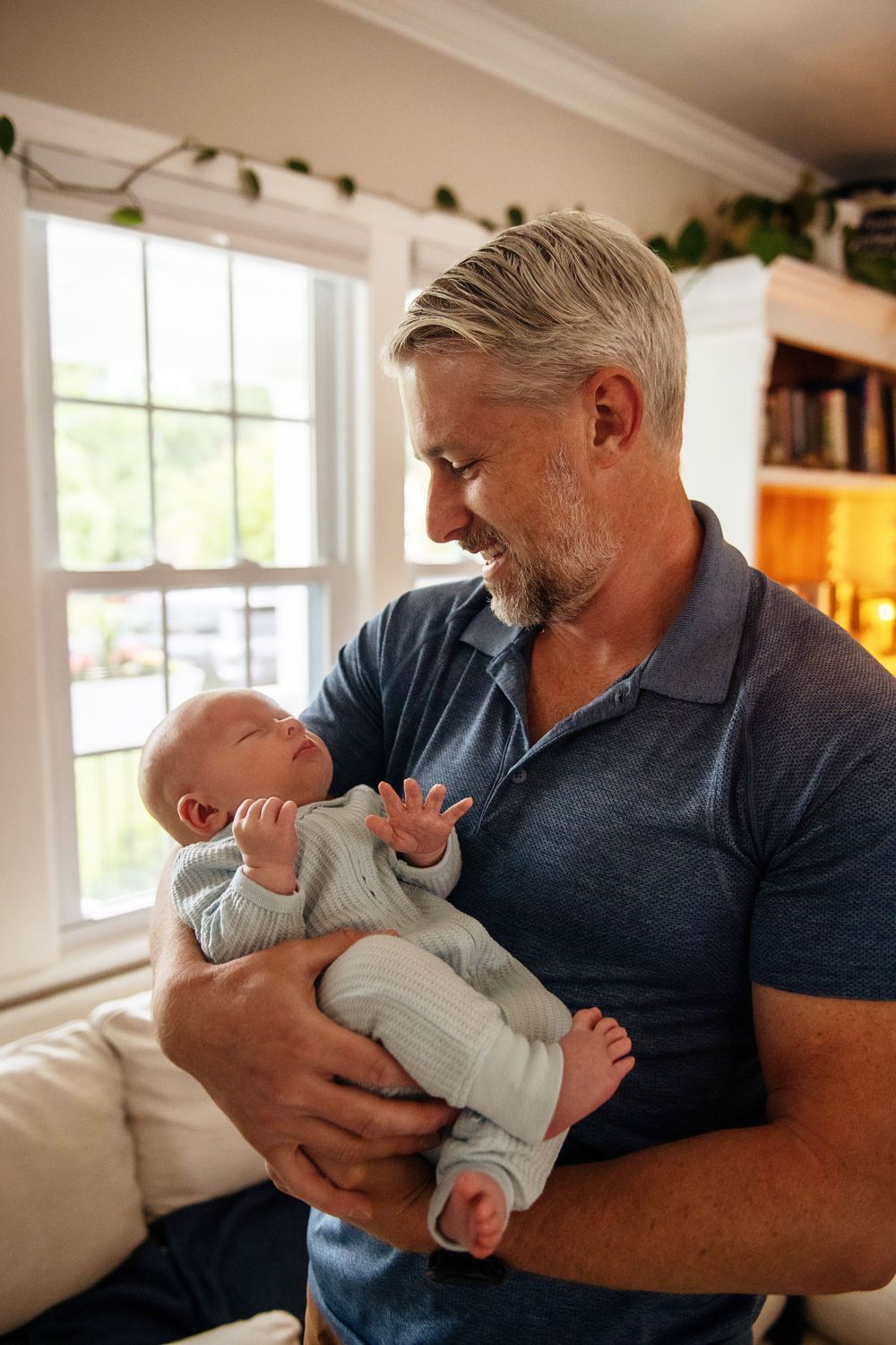 A loving caregiver holds a sleepy newborn baby while standing by a bright window in a cozy living room.