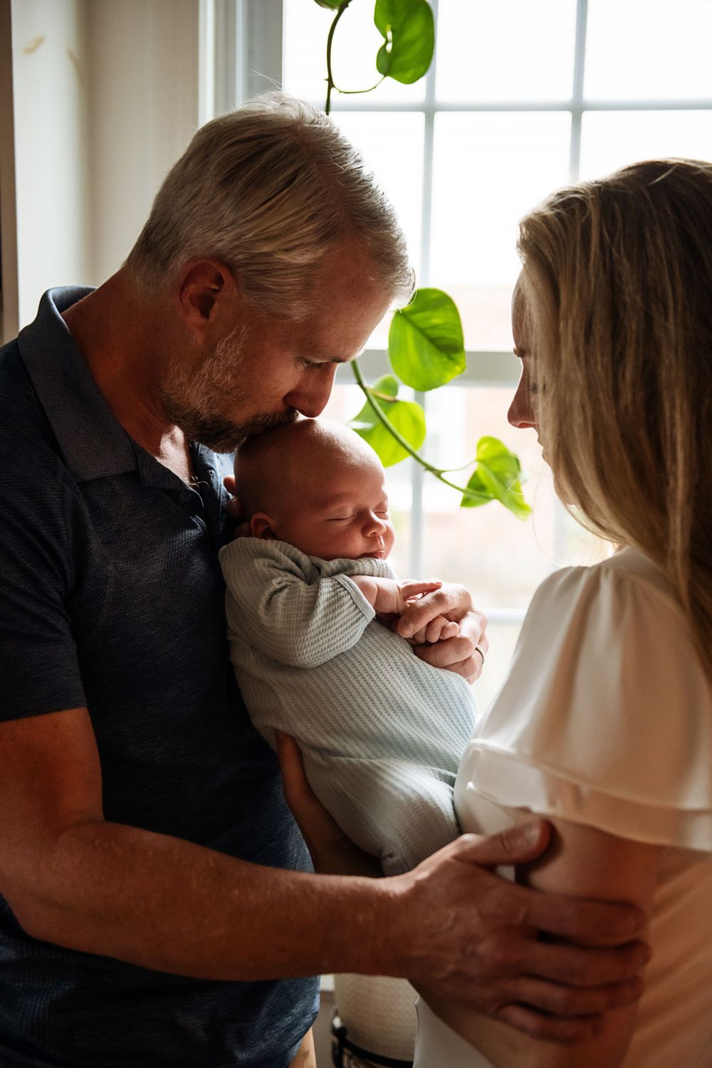 Couple embraces while holding their newborn baby near a sunny window with hanging plants.