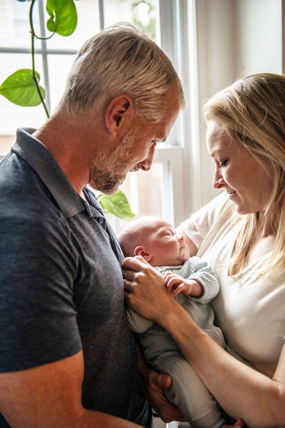 Intimate family moment as parents gaze lovingly at their newborn baby by a bright window.
