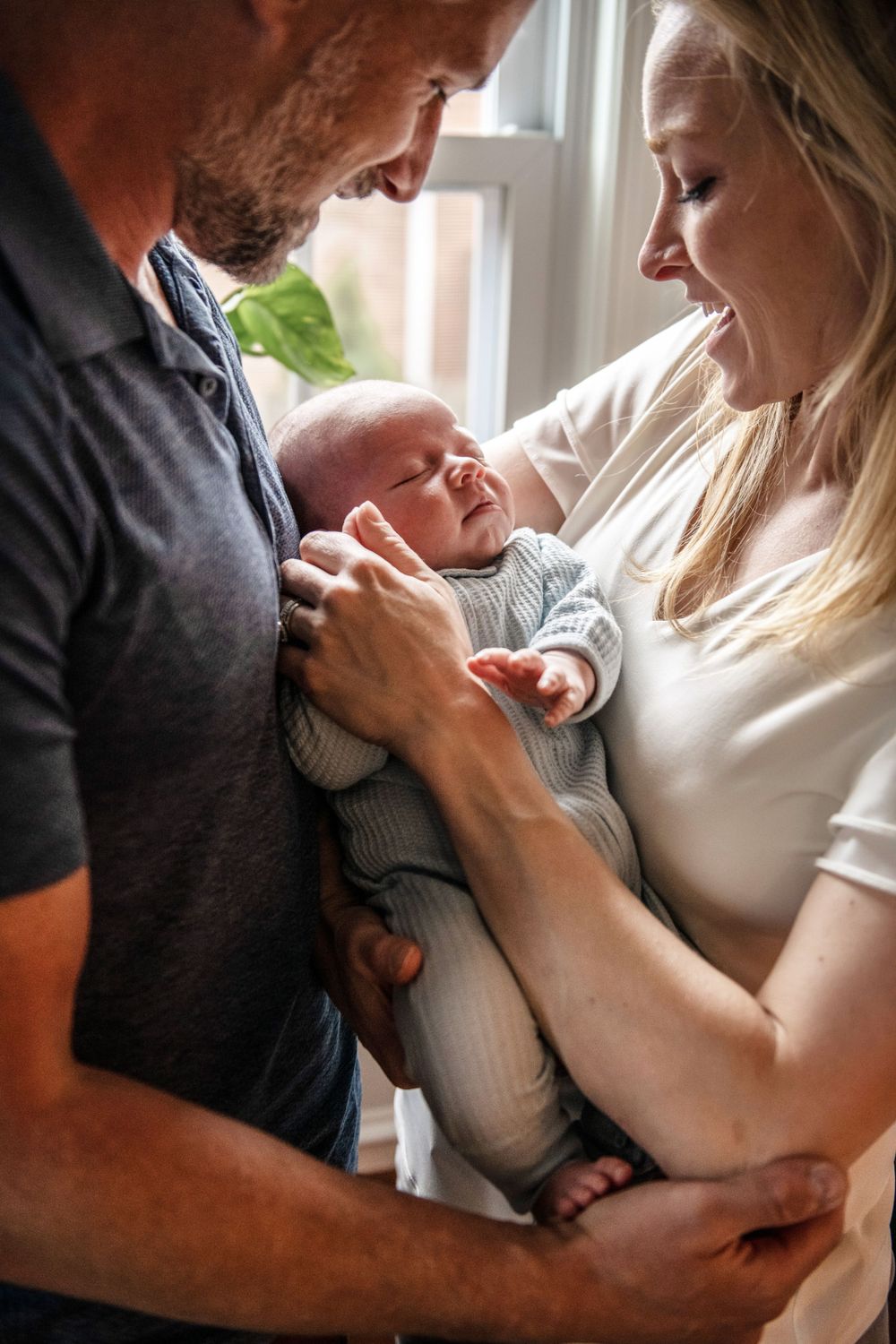 Parents share a joyful moment with their smiling newborn baby in soft window lighting.
