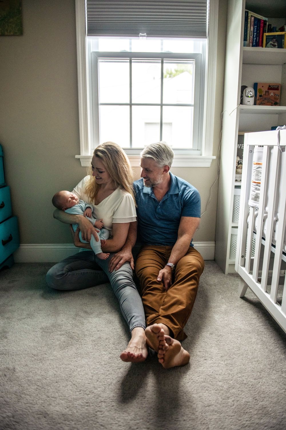 Family relaxing together on carpeted floor of nursery with white crib and window.